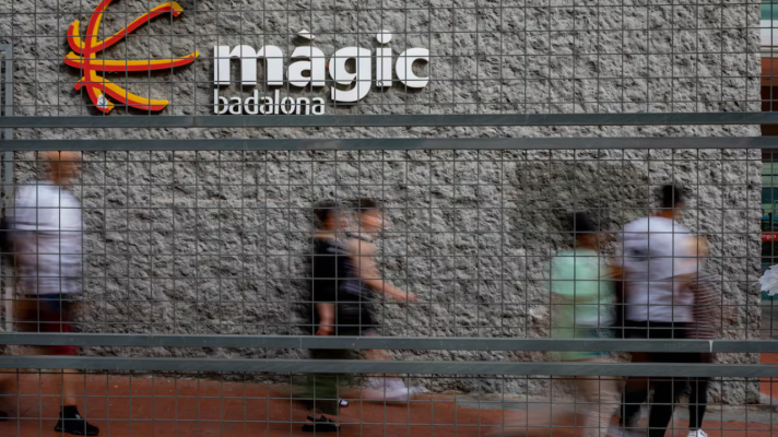 Fachada del centro comercial Màgic Badalona con su nombre en letras grandes y blancas, un logo y una valla metálica. Personas borrosas en primer plano.