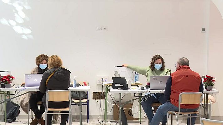 Cuatro personas con mascarillas, en mesas con ordenadores, atienden a otros dos durante la Navidad.  Se observan cables, dispositivos y papeles.