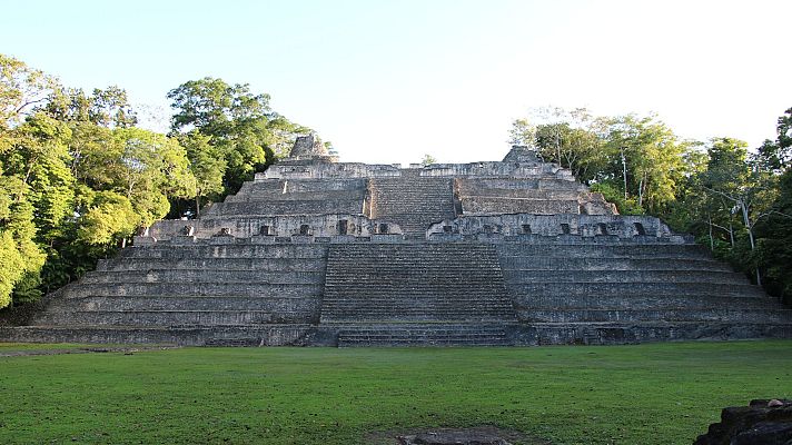 Pirámide maya escalonada con vegetación tropical circundante.  Muestra erosión y un área de césped al frente.  Imagen de un documental sobre la historia maya.