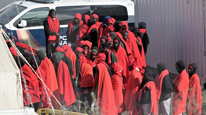 Numerosos migrantes, muchos con mantas rojas, en un centro de atención.  Se observan una tienda y un vehículo policial.
