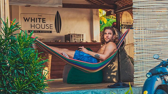 Hombre relajado en hamaca multicolor en terraza de hostel, con ventilador cercano y motocicleta azul.  Ambiente tropical.