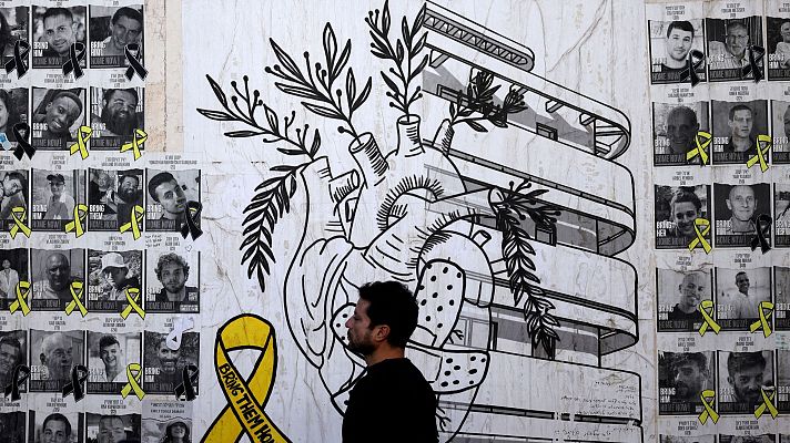 Mural en Tel Aviv con fotos de personas con lazos, una cinta amarilla con texto y un corazón con ramas.  Parece un memorial.