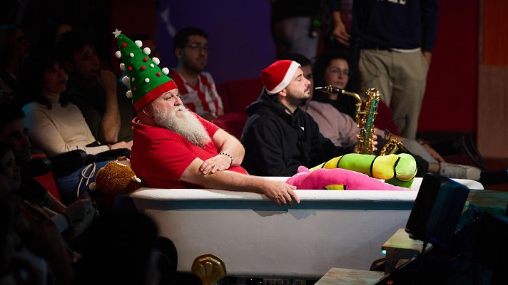 Papá Noel, con gorro de árbol, sentado en una bañera en un escenario, rodeado de peluches.  Un músico con gorro navideño toca el saxofón al fondo.
