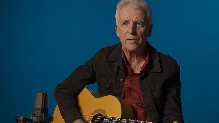 Kiko Veneno, con guitarra acústica, chaqueta negra y camisa granate, en imagen promocional.  Micrófono a la izquierda, fondo azul.