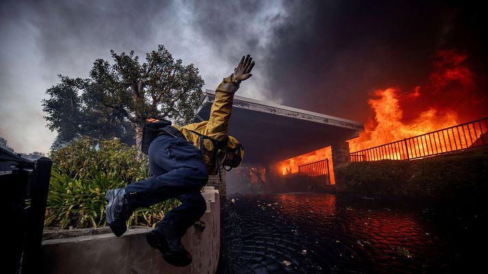 Bombero en acción saltando un muro durante un incendio.  Llamas intensas y humo denso. Suelo mojado.