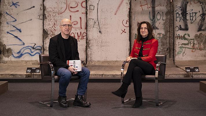 Dos personas sentadas ante un fragmento del Muro de Berlín con grafitis. Uno sostiene un libro titulado \"VICTOR\".  Señales indican \"no cámaras\" y \"no móviles\".