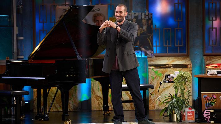 Humorista en plató de televisión, con chaqueta gris y pantalones oscuros, gesticula con las manos formando un corazón delante de un piano de cola.  El escenario incluye una mesa con objetos.