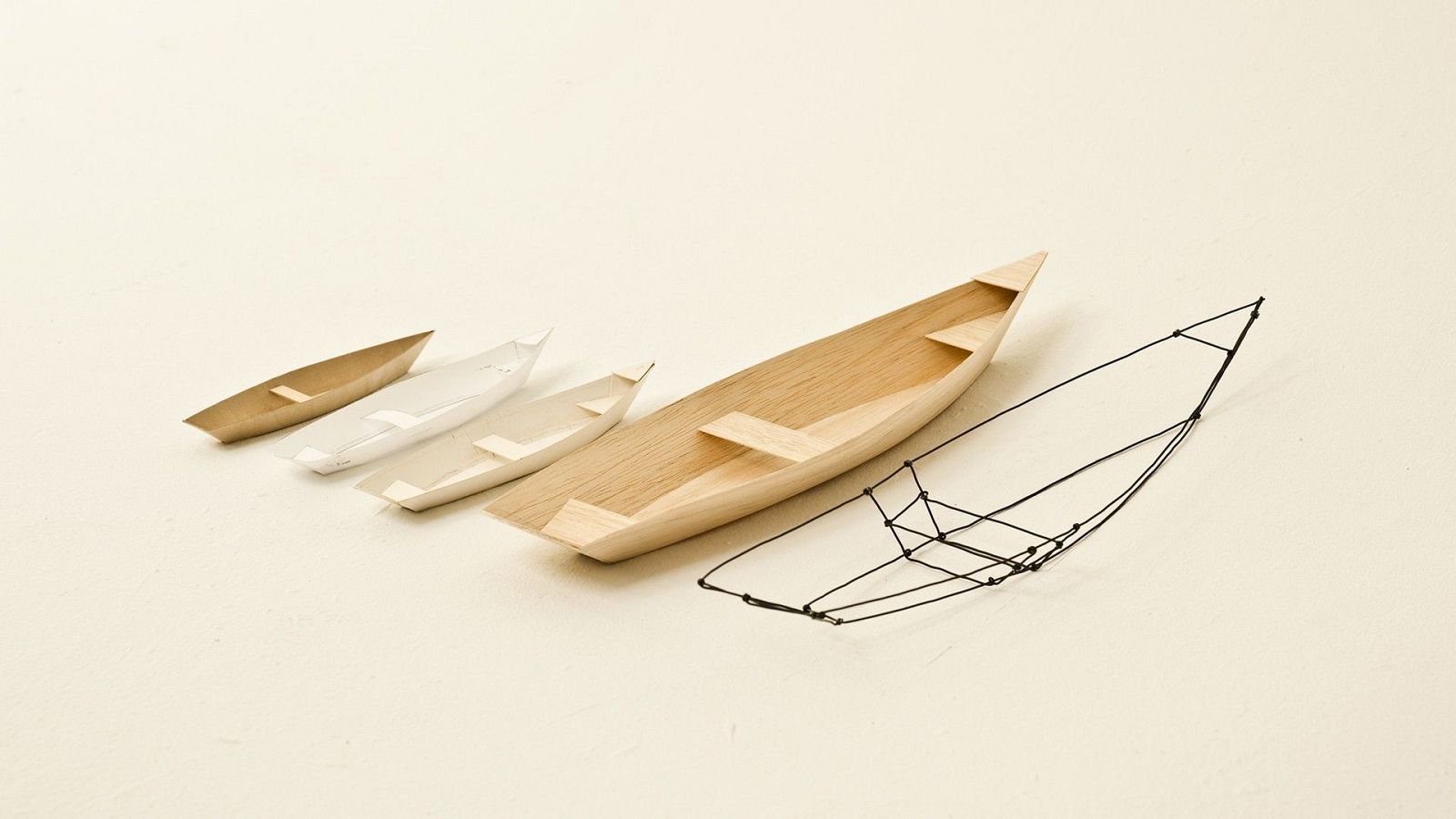 Cuatro maquetas de barcos de madera, de menor a mayor tamaño, junto a un armazón de alambre.  Posible representación de un proceso de diseño o construcción.
