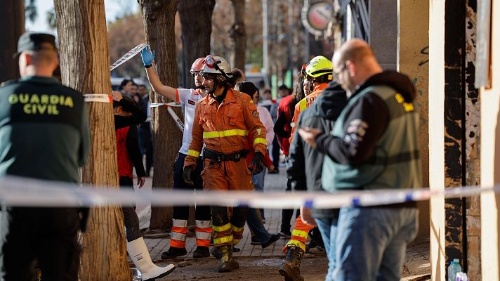 Intervención de bomberos en una emergencia.  Calle acordonada, suelo con escombros, civiles observando desde lejos.