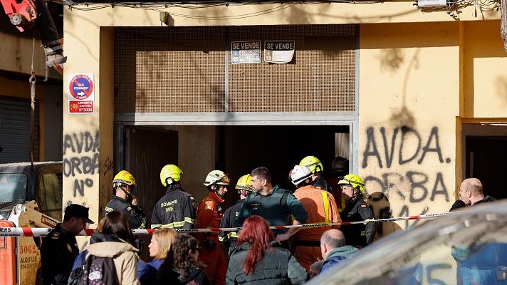 Emergencia en Benetússer: bomberos, policías y una excavadora trabajan en una zona acordonada frente a un edificio con carteles de \"SE VENDE\" y pintadas en la fachada.