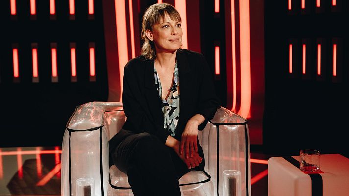 Mujer en traje negro y blusa floral sentada en sillón transparente en plató con iluminación roja.  Mesa baja blanca con vaso cerca. Ambiente de grabación o entrevista.