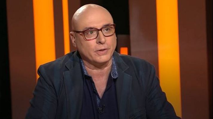 Pep Bras en un estudio de televisión, con fondo anaranjado, vestido con chaqueta oscura y camisa azul marino, aparentemente en una entrevista o conversación.