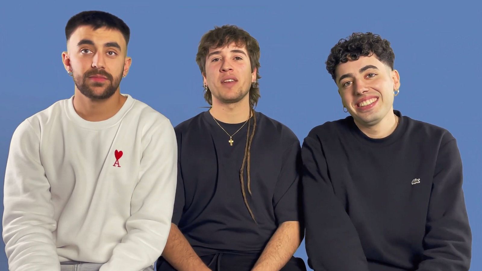 Tres jóvenes con ropa informal y pendientes posan sobre fondo azul.  Imagen promocional, posiblemente para un programa de radio o entrevista.