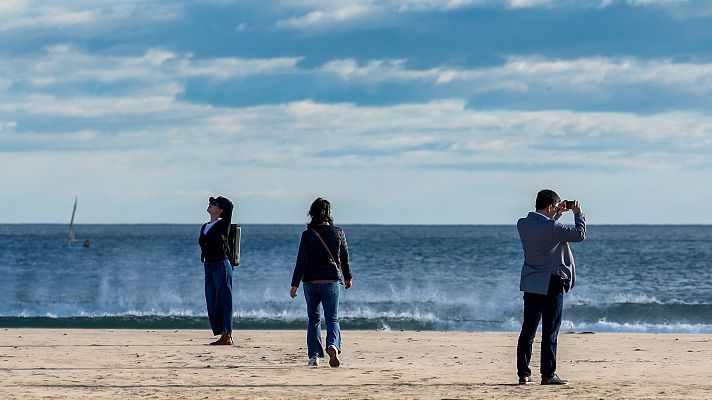 Día cálido en una playa valenciana: tres personas, una mirando al cielo, otra caminando y otra grabando con un móvil. Mar tranquilo, velero en el horizonte.