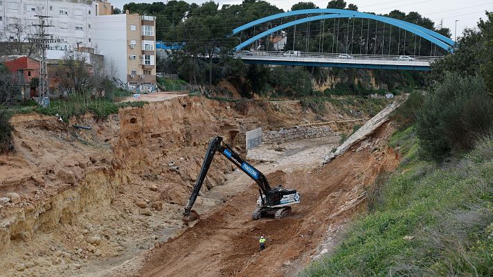 Excavadora trabajando en la reparación de un cauce fluvial erosionado tras una DANA.  Se observa un puente y una persona con chaleco de seguridad.