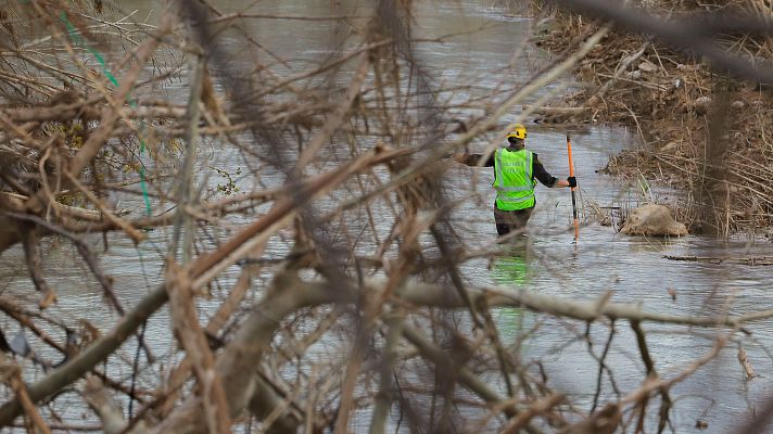 Miembro de equipo de búsqueda en río, con chaleco y casco amarillos, usa vara larga para inspeccionar el fondo.  Agua turbia, vegetación dañada.