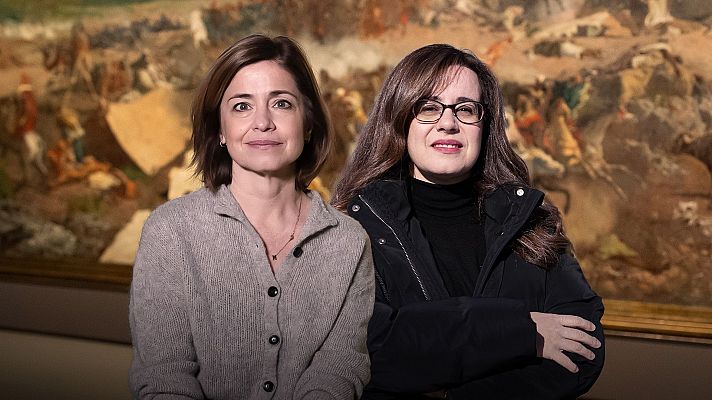 Dos mujeres, una con cárdigan claro y otra con chaqueta negra, posan ante una pintura de una escena de batalla con figuras a caballo y tiendas de campaña.