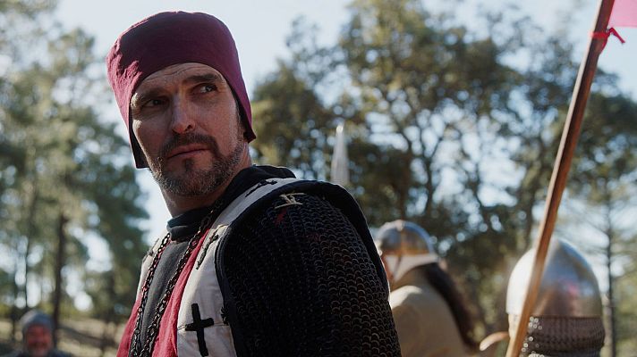 Soldado medieval con cota de malla y túnica cruzada, expresión seria, mirando a la derecha. Fondo con otros soldados y bandera roja.