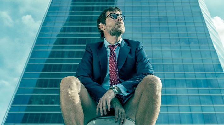 Hombre con americana, pantalones cortos y corbata roja con lunares blancos, sentado frente a un edificio de oficinas de cristal. Mira hacia arriba con expresión pensativa o desafiante.
