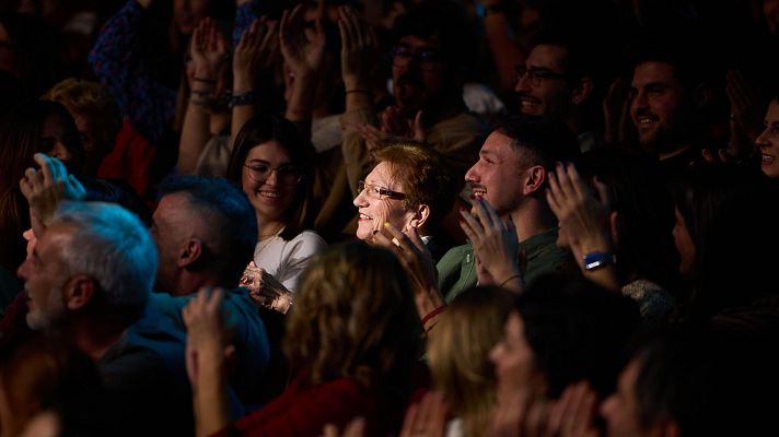 Público entusiasmado aplaudiendo en un evento.  Ambiente cálido y buena iluminación. Una mujer mayor con gafas disfruta del espectáculo.