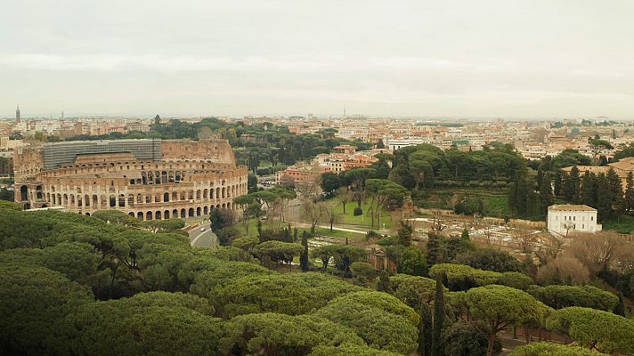 Vista panorámica del Coliseo romano, parcialmente en andamios, rodeado de vegetación y con la ciudad al fondo bajo un cielo nublado.