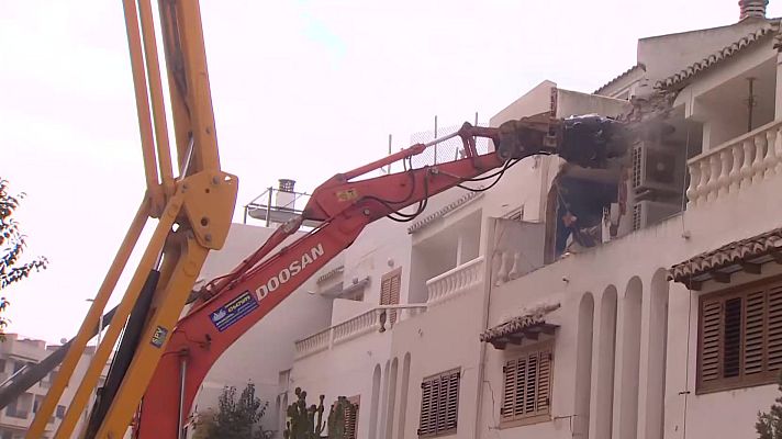 Excavadora demoliendo edificio de dos plantas; brazo extendido, generando polvo y escombros.  Detalles arquitectónicos visibles.