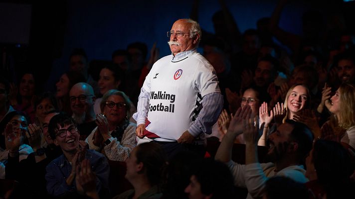 Hombre mayor con camiseta \"Walking Football\" es aplaudido por una audiencia en un escenario.  Momento de reconocimiento o presentación.
