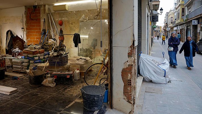 Interior de comercio en reconstrucción tras daños.  Materiales de construcción, escombros y suelo a medio instalar. Personas en la calle.