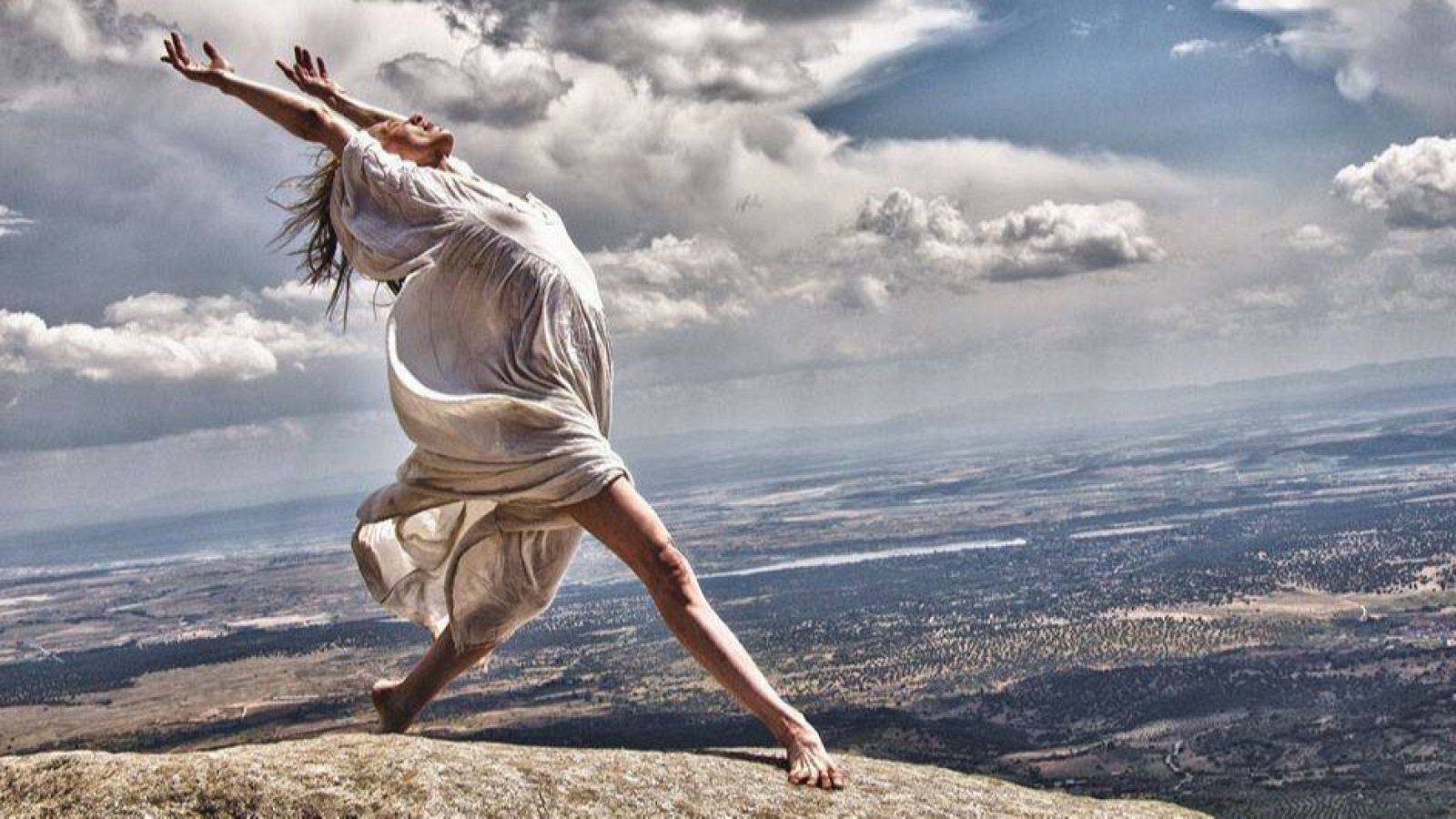 Imagen de una persona en una pose dinámica sobre una roca, con vestimenta blanca y un paisaje extenso de fondo.