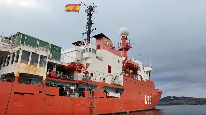 Barco de investigación rojo y blanco (A33) con bandera española, cerca de costa montañosa.  Vistas antenas, bote salvavidas y personal a bordo.