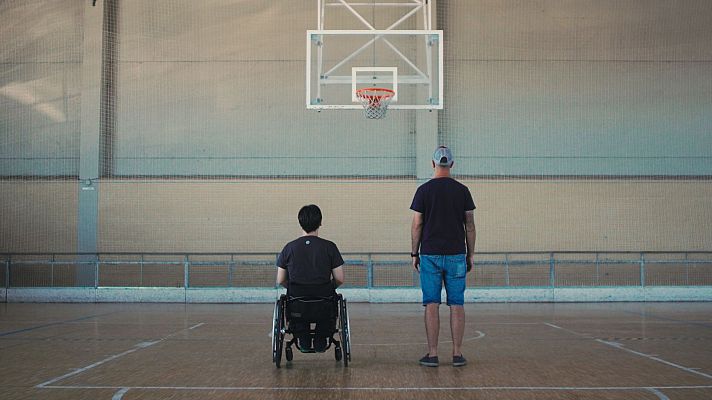 En un pabellón deportivo, dos personas, una en silla de ruedas, miran hacia una cancha de baloncesto. Atmósfera tranquila.