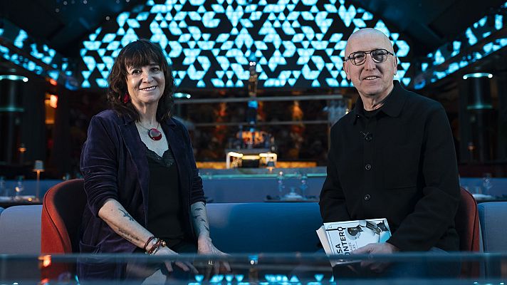 Dos personas, una mujer con chaqueta oscura y colgante rojo, y un hombre calvo con gafas sosteniendo un libro titulado \"LA COSTURERA\", sentadas en un sofá con fondo azul geométrico.