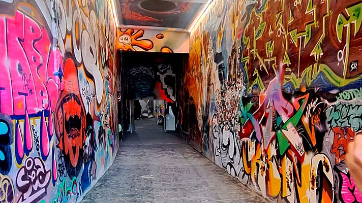 Pasillo estrecho con grafitis coloridos, figuras abstractas y personajes de dibujos animados. Suelo de cemento y una figura borrosa al fondo sugieren un espacio público.