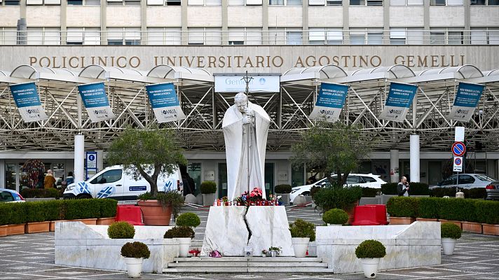 Ofrendas florales y velas ante una estatua del Papa Francisco en el Policlínico Gemelli de Roma.  Entrada del hospital visible al fondo.