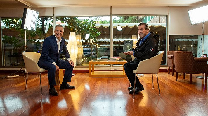 Entrevista grabada: dos hombres, uno en traje azul marino y otro con chaqueta oscura sosteniendo un libro titulado \"El hombre que buscaba la verdad en el mundo\", sentados frente a frente.