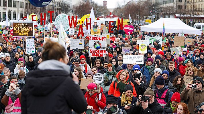 Multitud en manifestación alemana por el clima y temas sociales.  Pancartas con mensajes urgentes y una persona con micrófono dirigiéndose a los participantes.