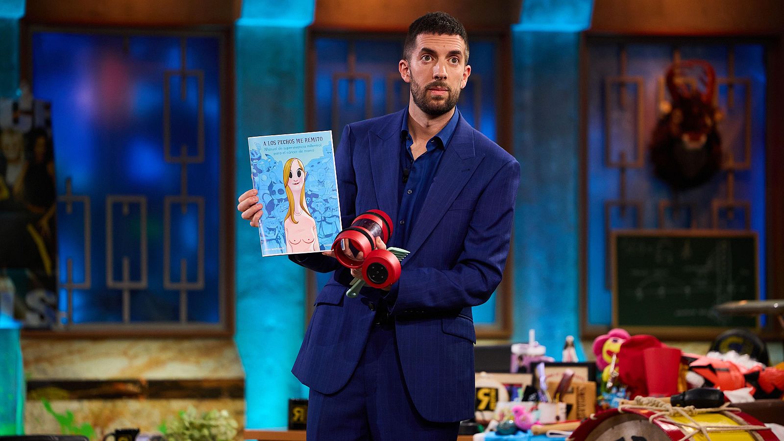 Presentador en plató con traje azul, mostrando un cómic con portada de mujer rubia y título "A GIRL PERIOD IN HA KUNITES", además de un objeto cilíndrico rojo.