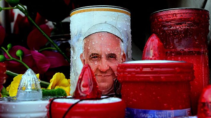 Lata con imagen del Papa Francisco, rodeada de velas (algunas encendidas) y flores, con gotas de agua visibles. Posible altar improvisado.