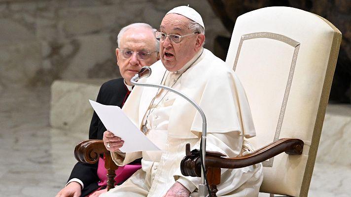 El Papa Francisco, sentado, lee un documento y habla durante una audiencia.  Un micrófono y otra persona con vestimenta religiosa están presentes.