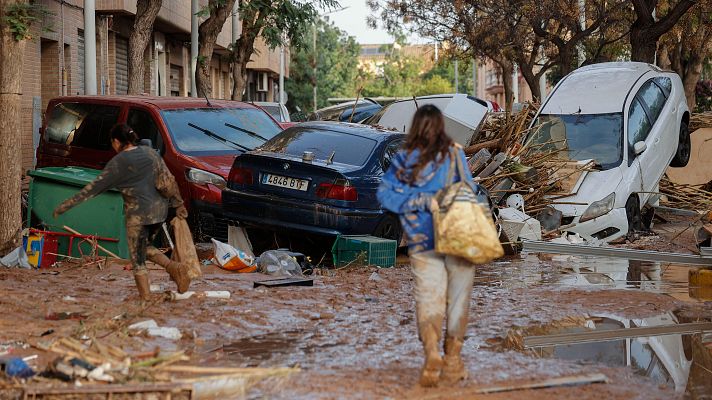 Calle inundada con barro y escombros tras una inundación. Coches dañados y personas intentando recuperar objetos.  Impacto devastador.
