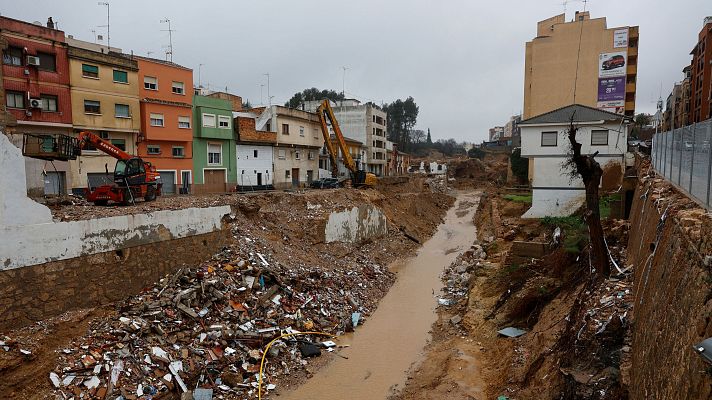 Desastre natural en zona urbana española: río de agua turbia entre escombros, edificios dañados y maquinaria pesada trabajando en la limpieza.