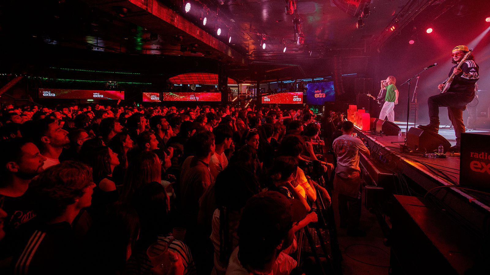 Intensa iluminación roja en concierto con músicos en escena; uno con guitarra, otro con micrófono.  Se observa el logo de Radio 3 Extra en pantallas y cartel.