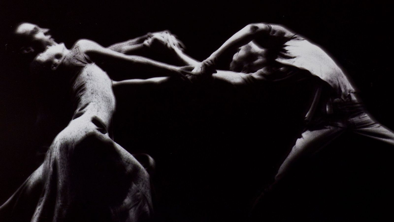 Imagen en blanco y negro de dos figuras en interacción física intensa, en un espacio oscuro con iluminación dramática.  Se aprecia tensión corporal y entrelazamiento de brazos y manos.