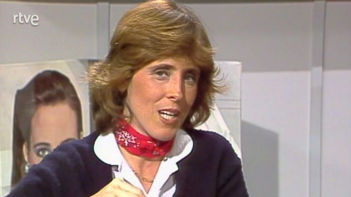 Mujer con pelo corto y rojizo, chaqueta oscura y pañuelo rojo, hablando en un programa de televisión. Fondo con imágenes borrosas y logo de RTVE. Aspecto granulado.