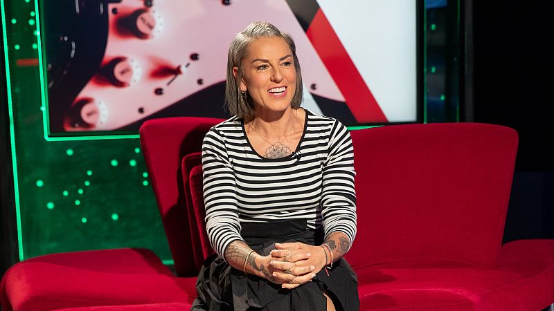 La Mari de Chambao, con el pelo corto y gris, sonríe durante una entrevista en un plató con sofá rojo y fondo verde. Lleva una camiseta a rayas y falda oscura.