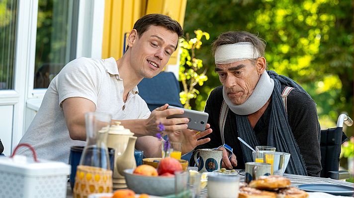 Dos hombres, uno lesionado (collarín y venda), comparten comida al aire libre mientras uno le muestra algo en su móvil.