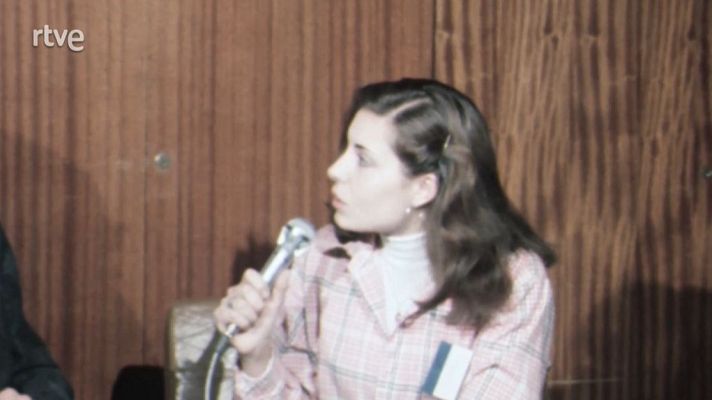 Mujer con camisa a cuadros y acreditación habla por micrófono en sala con paneles de madera.  Posiblemente una conferencia o evento.