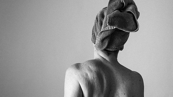 Espalda desnuda de una mujer mayor, con toalla en la cabeza, mostrando detalles de la piel que reflejan el envejecimiento. Imagen en blanco y negro.