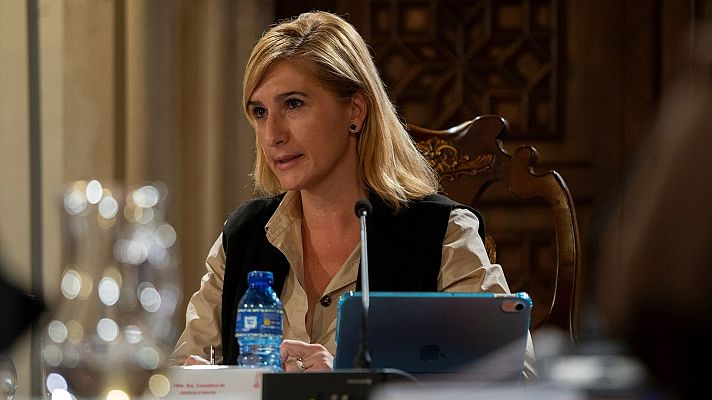Exconsellera sentada en mesa con micrófono, tablet y botella de agua. Entorno formal con silla de madera tallada y fondo ornamentado.