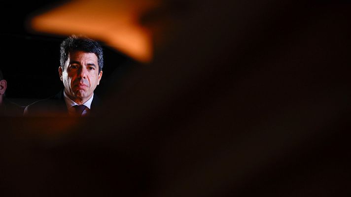 Carlos Mazón, con traje oscuro y corbata, en una foto oscura de interior con expresión seria.  A su izquierda, se vislumbra otra persona.